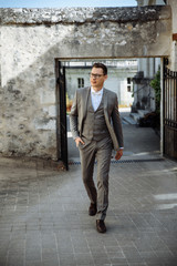 Obraz premium Perfect style. Handsome and very stylish young man in a suit and glasses walks along the street of the old town holding his hand in his pocket