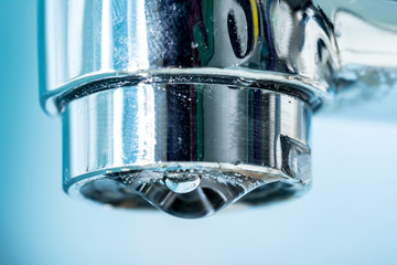 A drop of water hangs out of a modern chrome plated water tap faucet macro close-up.