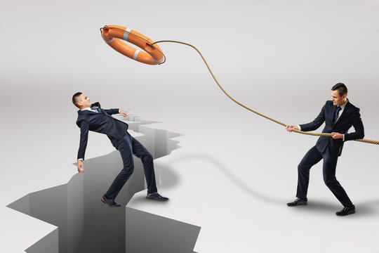 Businessman throwing orange lifebuoy to another businessman who is falling into earthquake crack on white background