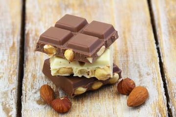 Pieces of white and milk chocolates with almonds and hazelnuts stacked up on wooden surface, closeup

