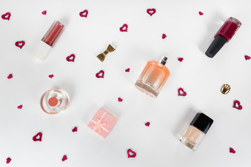 Cosmetic flat lay - perfume, nail polish, lipstick, small gift box, golden accessories on white background with tiny red hearts among them. Cosmetic sale, promotion, advertisement. 