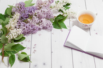 Beautiful branch of white and violet lilac flowers with opened pad, lying on the white wooden background with cup of tea, mock up perspective view with place for your text