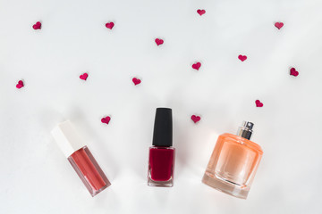 Cosmetic flat lay - perfume, nail polish, lipstick on white background with tiny red hearts among them. Cosmetic sale, promotion, advertisement. Valentine's day/ date night preparation.