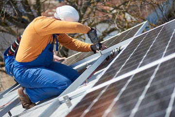 Male worker in protective helmet installing stand-alone solar photovoltaic panel system using screwdriver. Electrician mounting blue solar module on roof of modern house. Alternative energy concept.