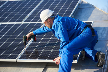 Male worker in blue suit and protective helmet installing solar photovoltaic panel system using...