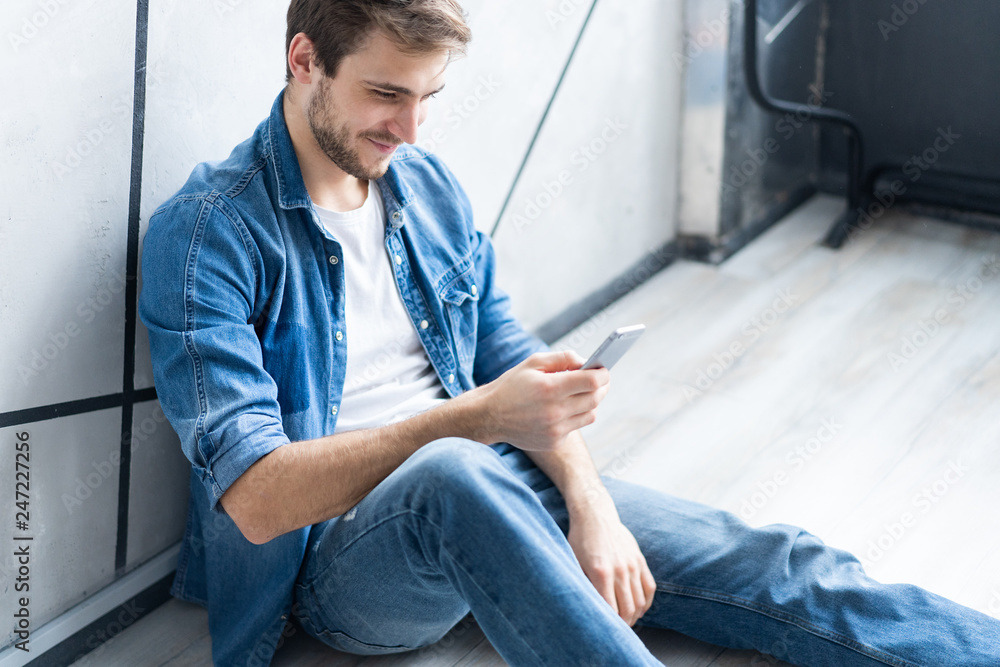 Wall mural Young smiling man sitting on floor while using smartphone against gray wall.