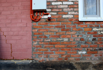 The wall of the house is made of red brick with a cement seam. Part of the wall is painted pink. The Foundation is black, cracked. Above hangs a metal box with red corrugated pipes. Next to the window