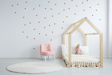 Pastel pink armchair next to wooden house shape bed with toy and blanket, copy space and golden...