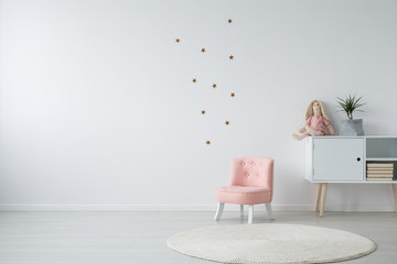 Pastel pink armchair next to wooden cabinet with books, toy and green plant in grey material pot,...