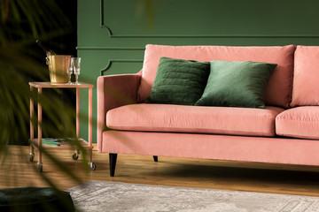 Emerald green pillows on pastel pink couch next to pink table with champagne glasses in trendy living room interior with dark green wall