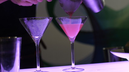  Barman in two beautiful glasses on high legs pours a cocktail from a shaker, glasses on a bar counter, a cocktail party, a competition of non-alcoholic cocktails