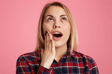 Headshot of surprised blonde woman focused upwards with astonished look, keeps jaw dropped from interest, has minimal make up, dressed in checkered shirt, isoalated over pink studio background