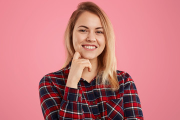 Portrait of good looking blonde female with gentle smile, keeps one hand under chin, dressed in checkered shirt, isolated over pink background. Photo of attractive woman models in studio alone