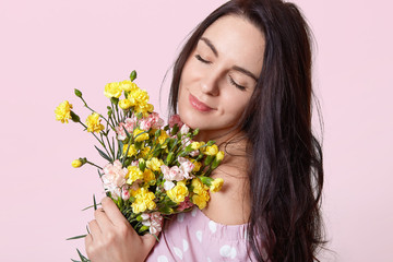 Obraz na płótnie Canvas Close up shot of dark haired woman with healthy skin, closes eyes with enjoyment, holds pretty flowers closely, poses over rosy background, feels pleased after date with boyfriend. Women concept