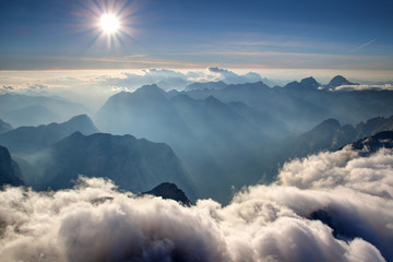 View from Triglav summit with blue ridges of Julijske Alpe range in sunshine, white low heap clouds, hazy Trenta Soca Isonzo valley and Bavski Grintavec peak, Triglav National Park Slovenia Europe