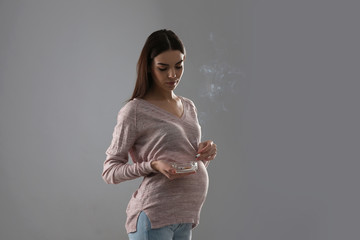 Young pregnant woman smoking cigarette on grey background
