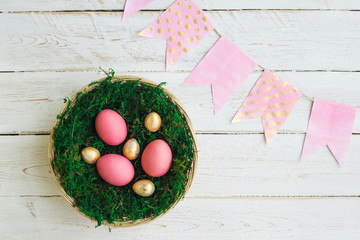 Easter holiday. Pink and gold colored Easter eggs lie in a basket with moss on a white wooden background. Selective focus