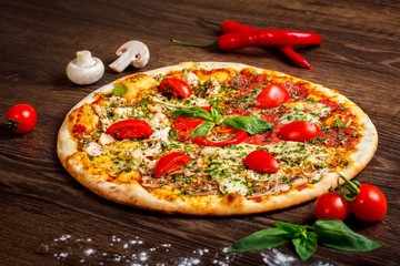 Pizza Italian with melted cheese, red cherry tomatoes, chicken, hen and fresh basil green leaves on a brown table decorated by mushrooms, red sweet pepper and cherry tomatoes