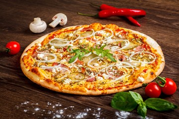 Pizza Italian with melted cheese, mozzarella, ham, pickled cucumbers, onion and fresh arugula leaves on a brown table decorated by mushrooms, red sweet pepper and cherry tomatoes