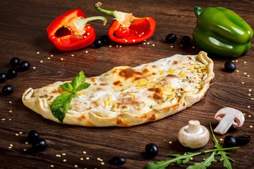Calzone Italian pizza with melted cheese, tomatoes, fresh green herb basil leaves on a brown table decorated by mushrooms, black olives, red and green pepper and arugula leaves