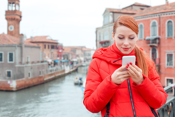 Cute smiling Latina woman using smart phone in Venice Italy. Woman in red coat winter clothing.