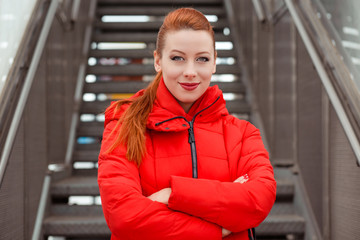 Pretty young confident redhead woman smiling standing near stairs in a urban background, hands crossed wearing red coat winter clothes