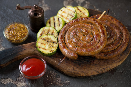Rustic wooden serving board with grilled coiled sausages, zucchini slices and dipping sauces, studio shot