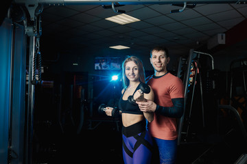 Obraz na płótnie Canvas Active beautiful fitness model girl trains biceps with dumbbells together with a trainer in the gym.