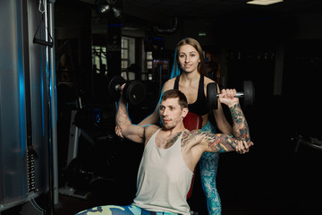 Fototapeta na wymiar Active beautiful fitness model girl helps to train with dumbbells while sitting to her boyfriend in the gym.