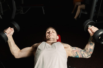 Handsome athletic man trains pectoral muscles by breeding dumbbells while lying in the gym.