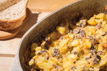 braised potatoes with mushrooms in a large cast-iron pan. on a wooden table sliced bread. view from above
