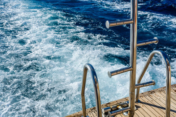 Metal staircase for descent into the water and wave trace with white foam on a water surface behind...
