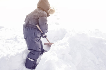little boy in overalls clears snow with a shovel. Sunny winter day.