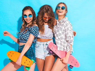 Three young stylish smiling beautiful girls with colorful penny skateboards. Woman in summer hipster checkered shirt clothes posing near blue wall in studio. Positive models having fun and going crazy