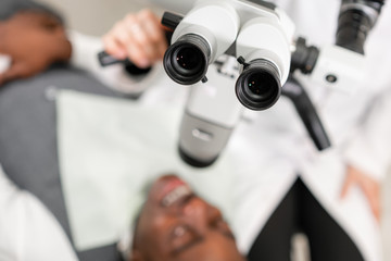 Eyepiece of microscope in foreground. Young African American male patient at chair at dental clinic. Medicine, health, stomatology concept. dentist conducts inspection and concludes