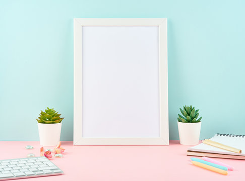 Mockup with blank white frame, alarm, notepad, keyboard on pink table against blue wall with copy space. Modern bright office desktop