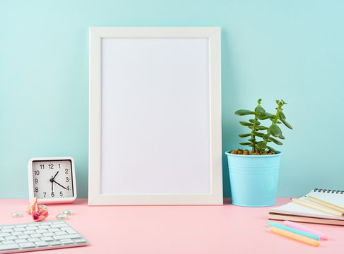 Mockup with blank white frame, alarm, notepad, cup of coffee on pink table against blue wall with copy space.