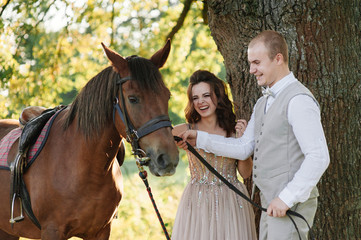 Obraz premium Wedding at sunset. Couple is sitting on a bench under a tree. Beige dress with sparkles. Light suit with a bow tie. The bride and groom embrace and kiss.