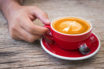 man hand hold red cup of coffee with milk making for flower picture on plate and spoon put on bark wooden table