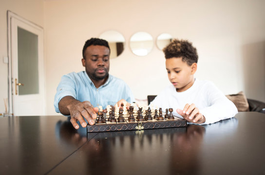 Cute african american son playing chess with his black father. The son teaches his father to play chess. Father teaches his son to play chess. Hobby for children. 