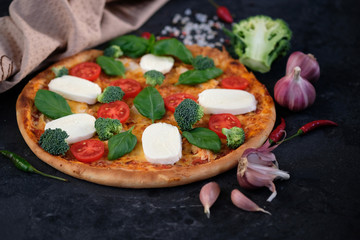 Italian pizza with Mozzarella cheese, tomatoes, broccoli, Spices and fresh basil. Pizza and kitchen napkin near, on black stone background