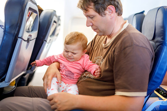 Young Sad Tired Father And His Crying Baby Daughter During Flight On Airplane Going On Vacations. Dad Holding Baby Girl On Arm. Air Travel With Baby, Child And Family Concept. Baby Wiht Earache.