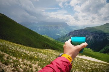 First-person view of a man's hand holding a plastic cup of tea against a plateau of green hills and...