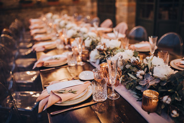 in backyard of villa in Tuscany there is banquet wooden table decorated with cotton and eucalyptus compositions, glasses, burning candles and plates are placed on table, evening