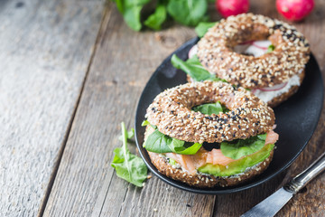 Bagels with cream cheese avocado, fish, arugula and radish on old wooden table. Healthy breakfast food.