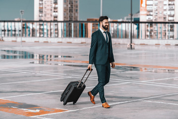 Fototapeta premium Full length of smiling businessman in formal wear with luggage walking on the parking. Business trip concept.