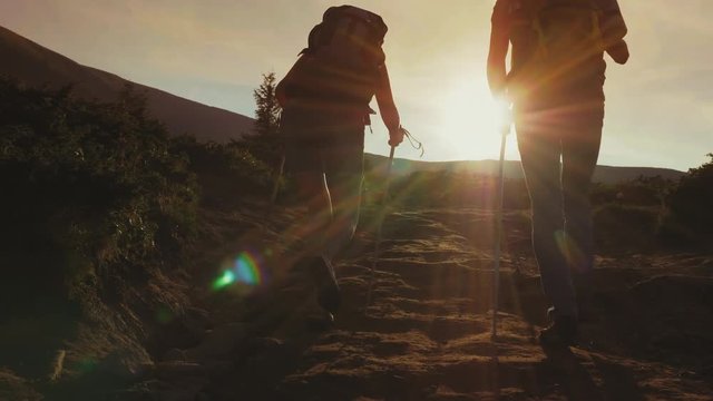 Two travelers with backpacks go forward along the mountain path towards the sun