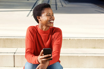smiling young african american woman sitting outside with mobile phone