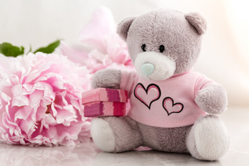 Little grey teddy bear sitting with a gift box and pink peonies behind him. Romantic gift ideas, presents for children.
