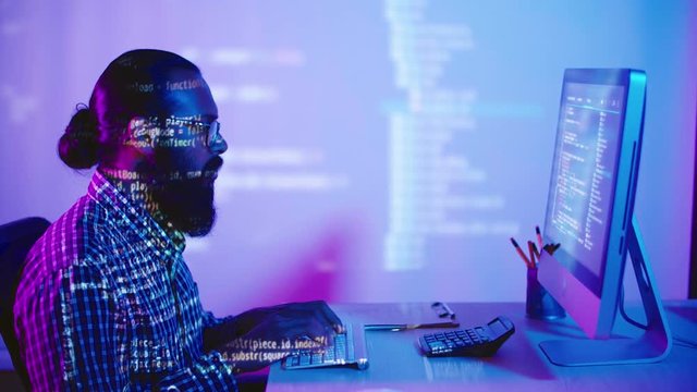 Lockdown of young Indian programmer with beard sitting at computer and typing code of new software, while computer screen light and code letters reflecting on him and on walls